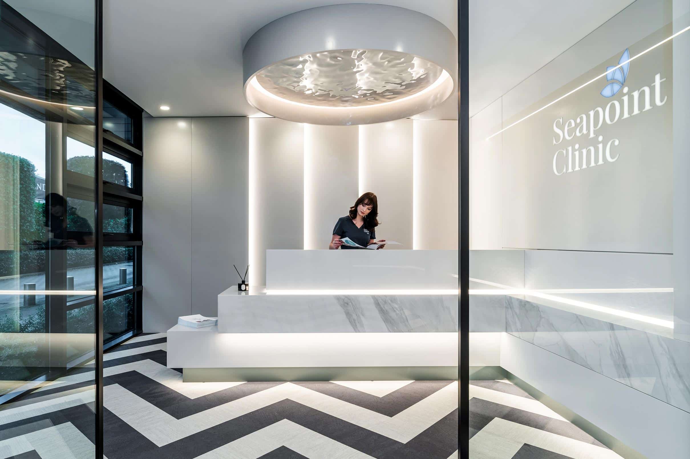 A modern clinic reception area with radian geometric flooring, a circular ceiling feature, and a receptionist standing behind a white marble desk. Seapoint Clinic is displayed on a wall with a butterfly logo.